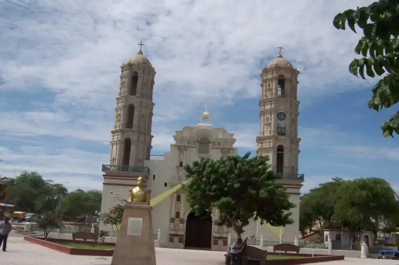 Catedral de Sechura, con dos torres blancas, junto a una plaza con árboles bajo un cielo azul