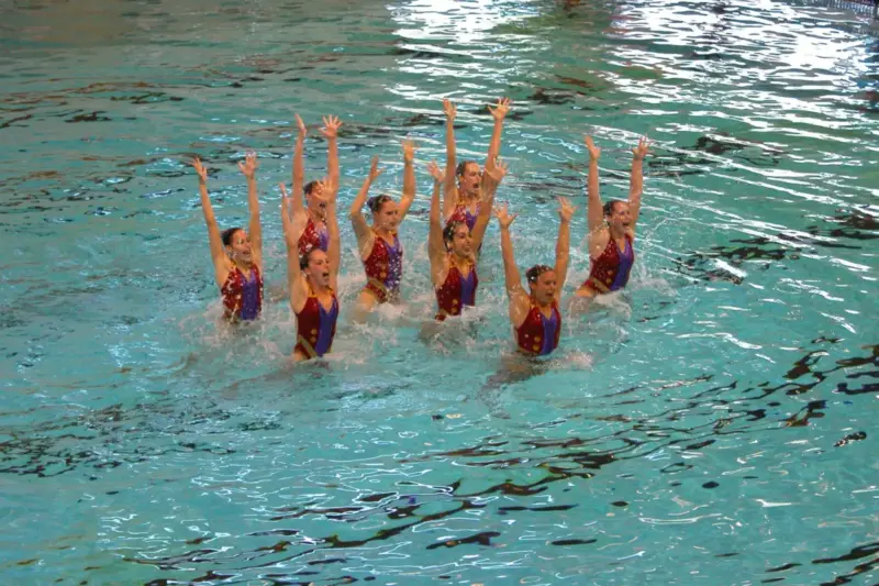 Un equipo de natación artística levanta los brazos a la vez en el agua de una piscina