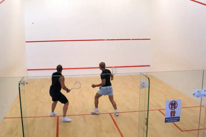 Dos jugadores de espaldas disputan un partido en una pista de squash de paredes blancas y suelo de madera