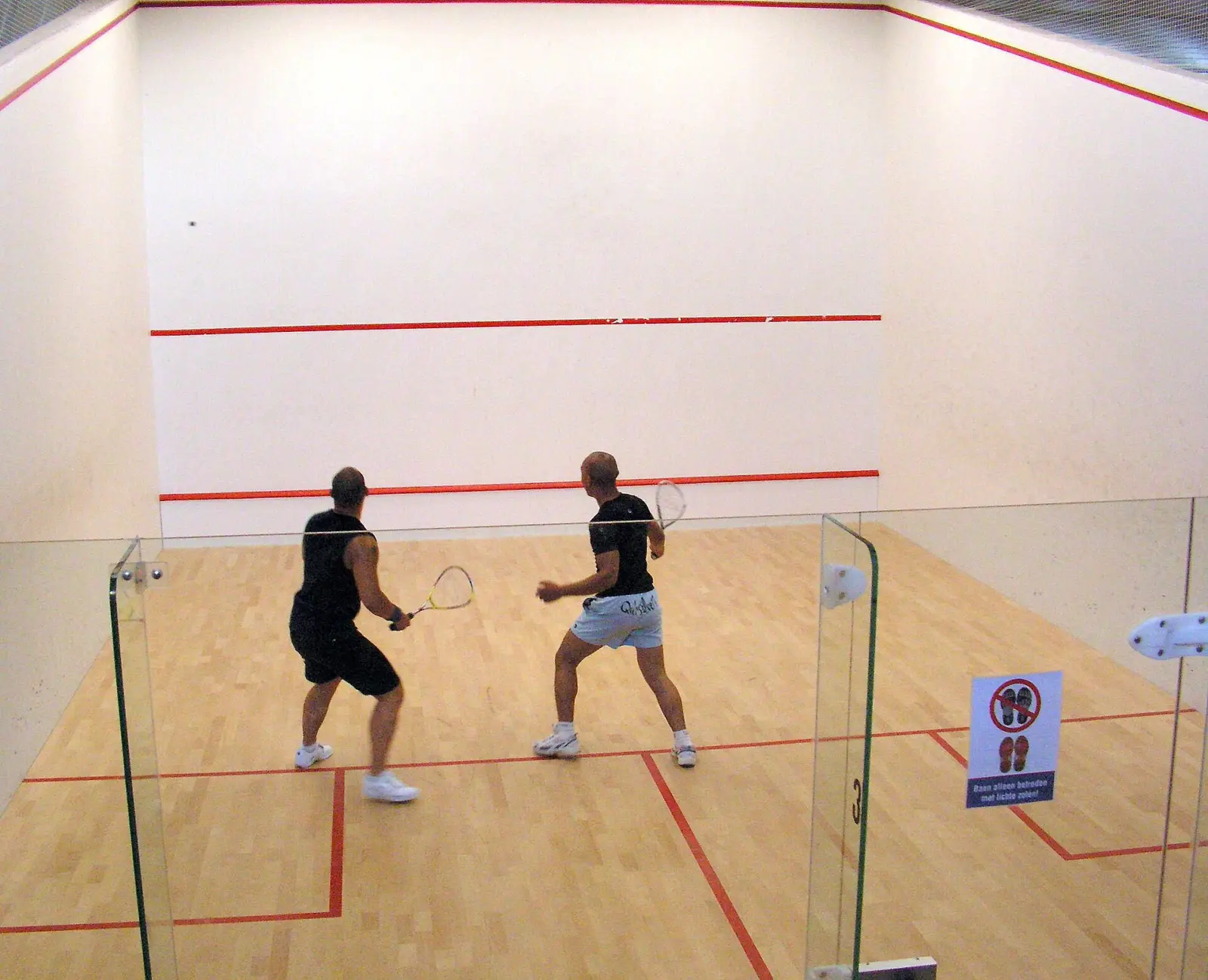 Dos jugadores de espaldas disputan un partido en una pista de squash de paredes blancas y suelo de madera