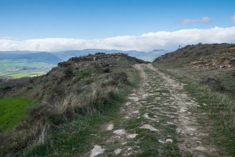 Antigua calzada romana de piedra que avanza por una loma herbosa con montañas y nubes al fondo
