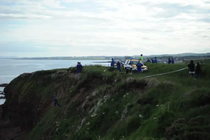 Un rescatista desciende con cuerdas por una ladera junto al mar mientras un equipo espera arriba
