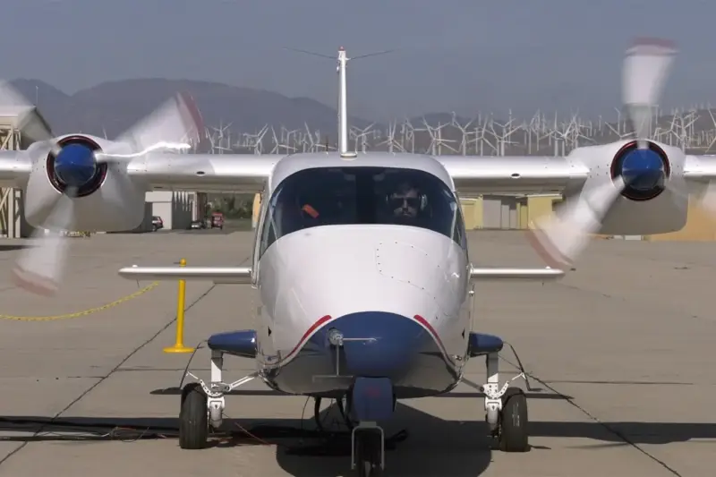 Avión eléctrico de hélices en la pista de un aeródromo, con aerogeneradores al fondo