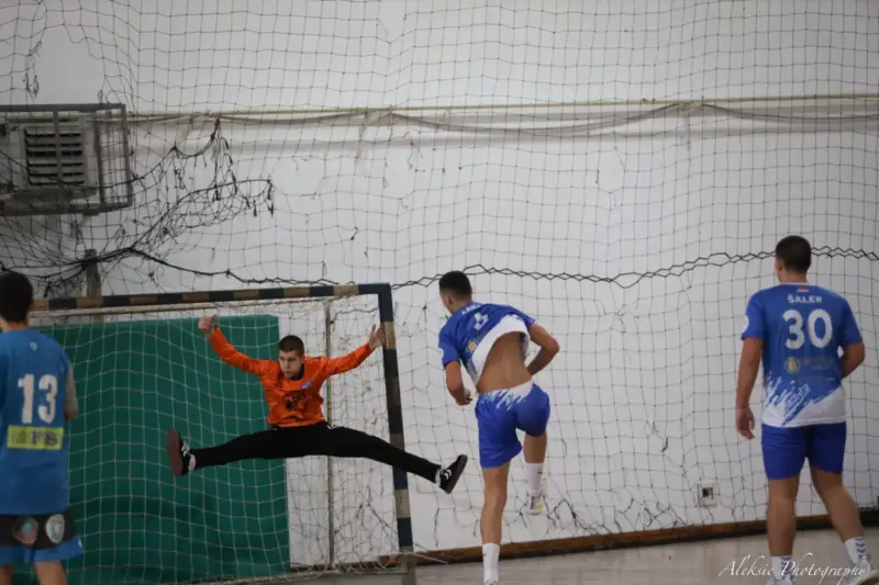 Portero de balonmano parando un lanzamiento en una pista cubierta durante un partido nocturno.