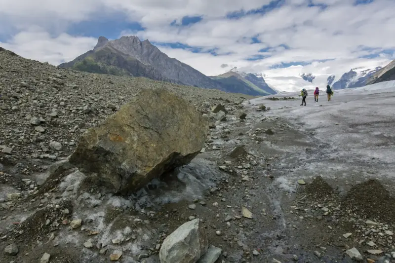 Barril de madera deformado entre rocas y hielo de un glaciar alpino, con un excursionista empujando una carretilla.