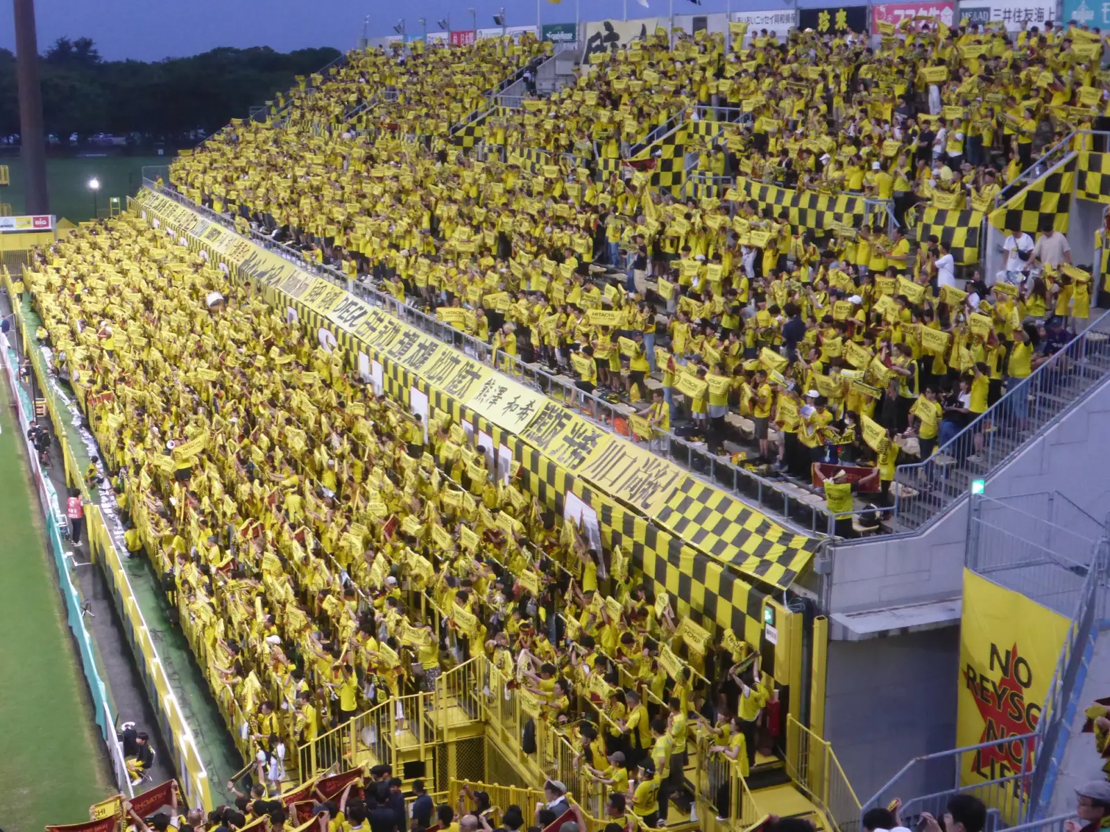 Aficionados en la grada de un estadio de fútbol durante un partido de noche, con la mayoría mirando el terreno de juego.