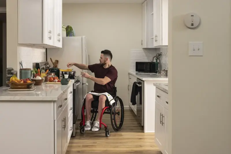 Hombre sentado en silla de ruedas frente a un ordenador en una cocina iluminada por la tarde.