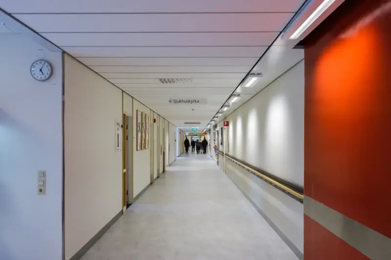 Mujer sentada en la sala de espera de un hospital, con una carpeta sobre las rodillas, junto a una ventana con luz natural.