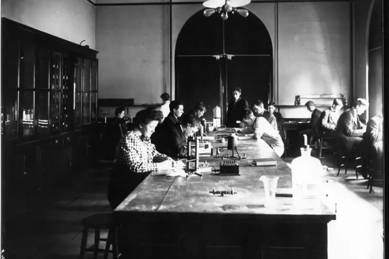 Estudiante tomando apuntes en un aula universitaria con una pizarra y estanterías de libros al fondo.