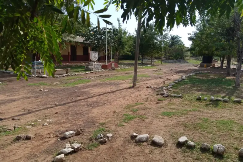 Niñas con uniforme caminando hacia una escuela rural mientras una mujer las observa desde la entrada.