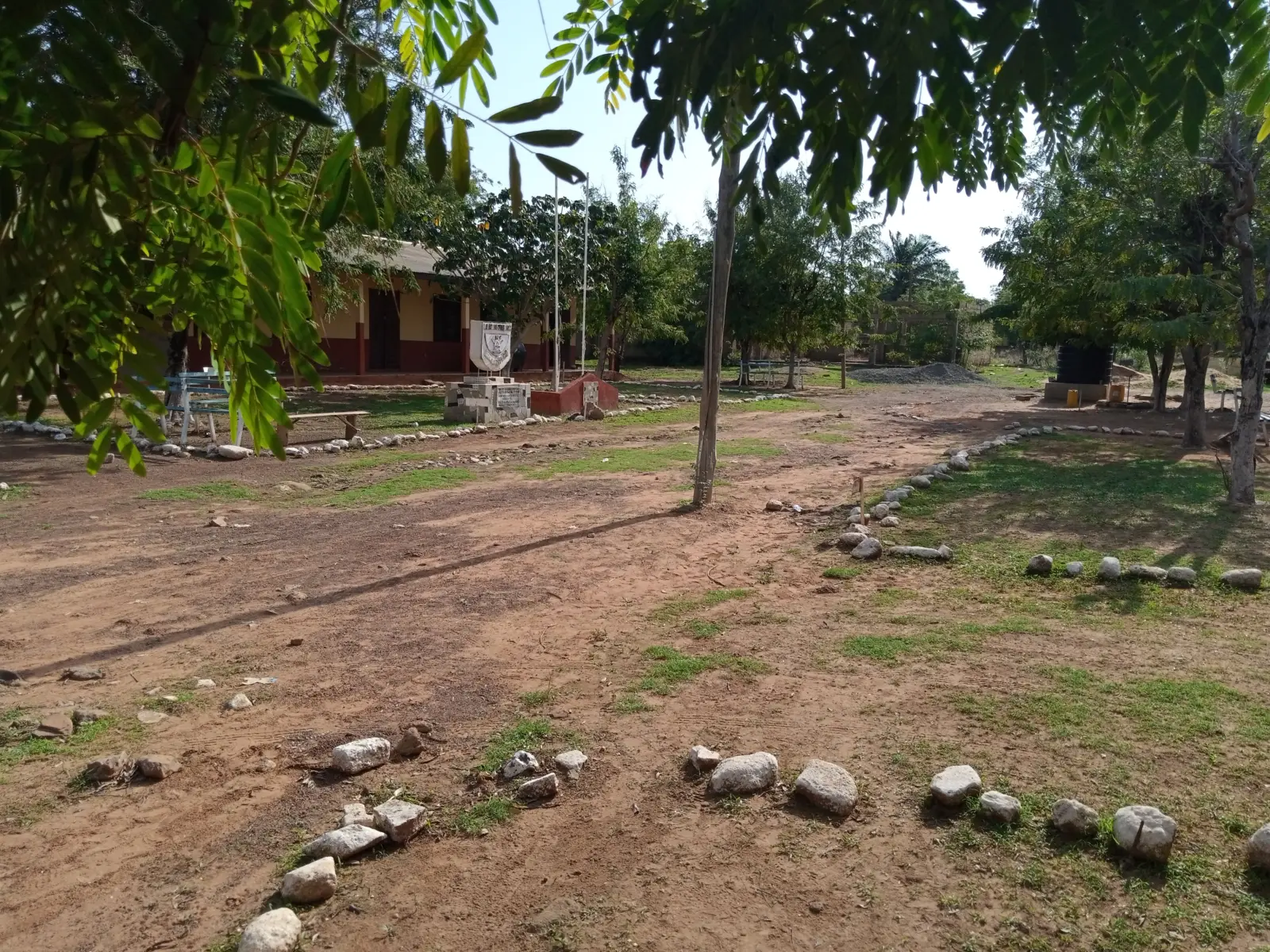 Niñas con uniforme caminando hacia una escuela rural mientras una mujer las observa desde la entrada.