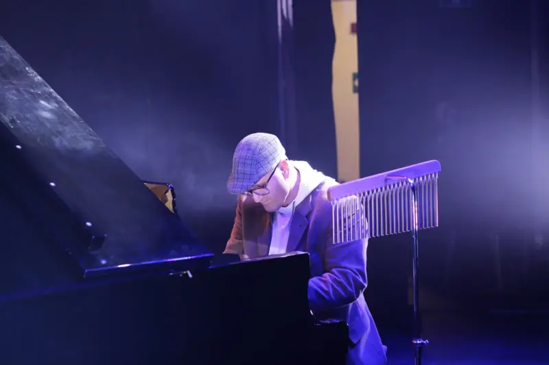 Escenario vacío con un piano de cola iluminado por focos cálidos, cortinas azules y confeti en el aire.