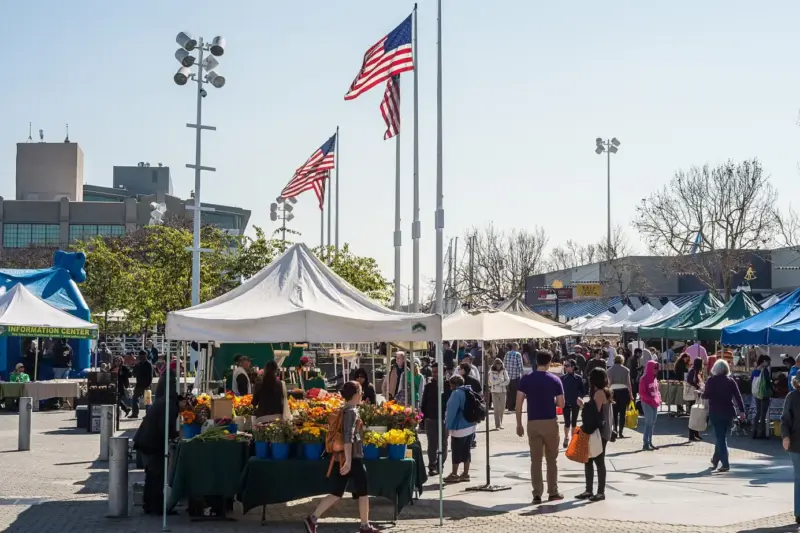 Puesto de productos frescos en un mercado al aire libre, con cajas de frutas y verduras y personas al fondo.