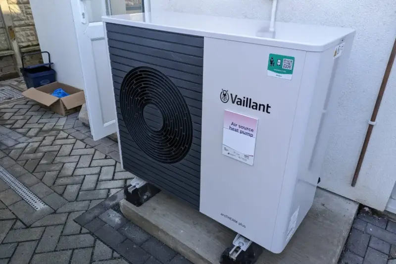 Unidad exterior de una bomba de calor junto a la fachada de una vivienda, con paneles solares en el tejado.