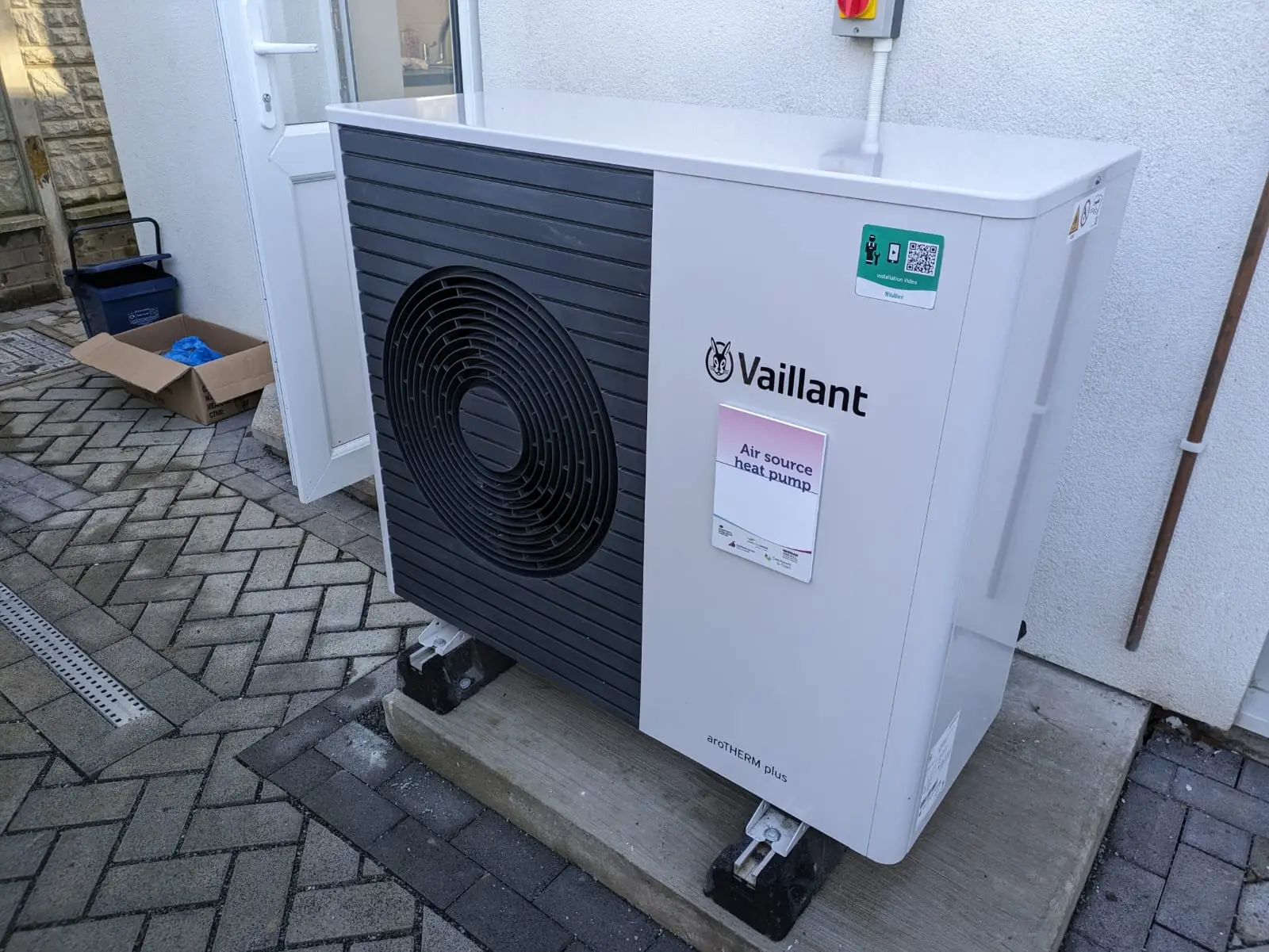 Unidad exterior de una bomba de calor junto a la fachada de una vivienda, con paneles solares en el tejado.