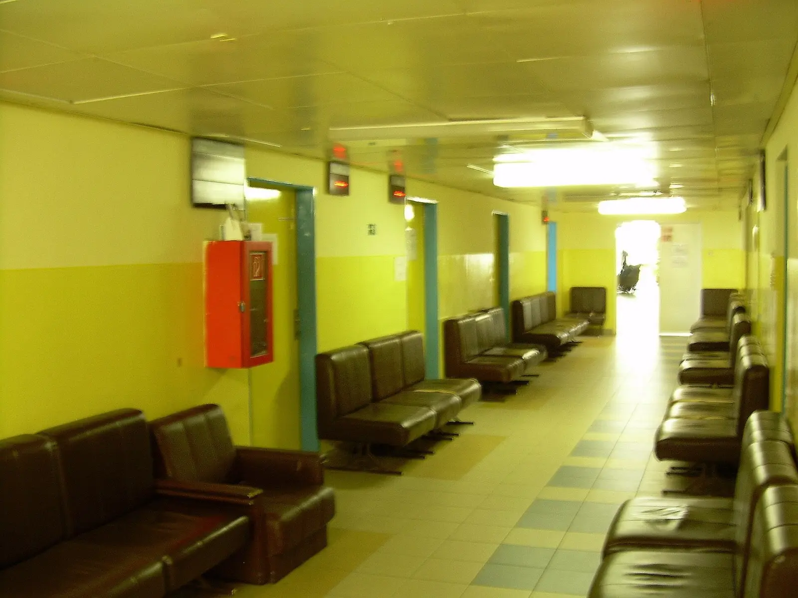 Sala de espera de un centro de salud con luz natural, un papel de cita sobre un banco de madera y una persona mayor al fondo.